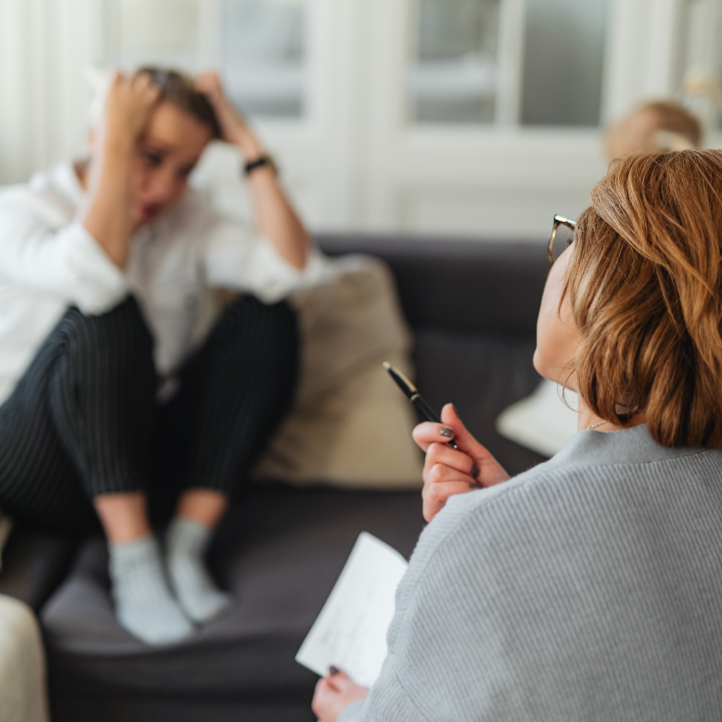 Therapist holding a pen during a counseling session while a client sits on a couch in the background with hands on their head